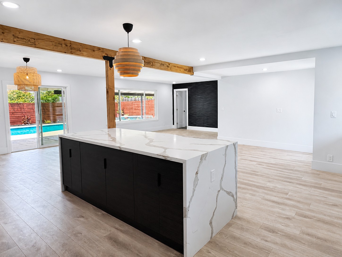 Open great room — waterfall island, wood beam, twin pendants, slider to the pool, 3D wave accent wall
